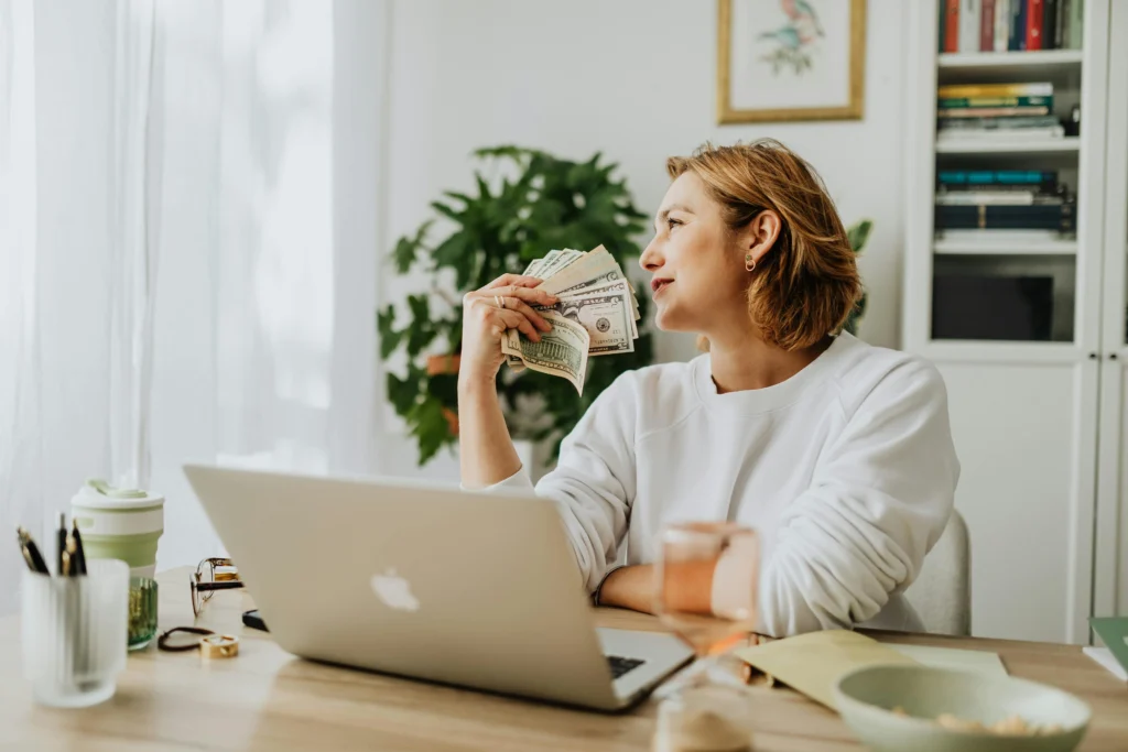 "A woman sitting at a desk with a laptop, holding cash and smiling, representing financial independence through side hustles."